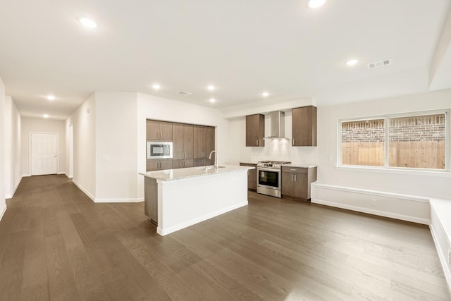 kitchen with light stone countertops, wall chimney exhaust hood, an island with sink, wood-type flooring, and appliances with stainless steel finishes