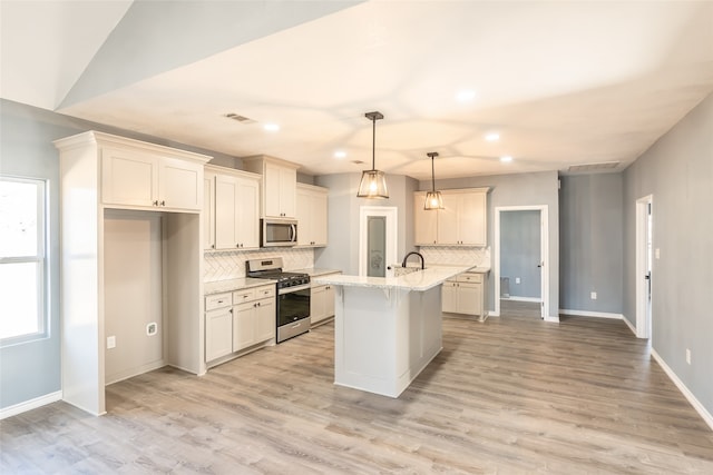 kitchen with light wood-type flooring, a kitchen island with sink, appliances with stainless steel finishes, decorative light fixtures, and light stone countertops