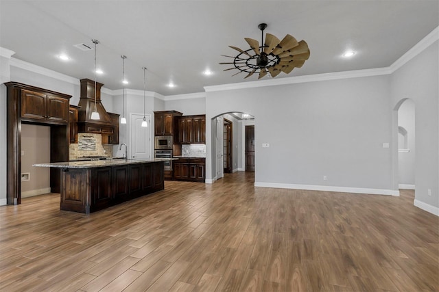 kitchen with custom exhaust hood, decorative light fixtures, dark brown cabinets, light stone countertops, and a kitchen island with sink