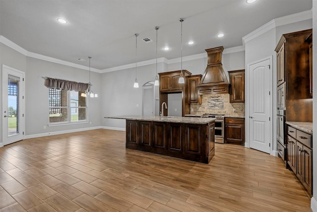 kitchen with appliances with stainless steel finishes, decorative backsplash, custom exhaust hood, hanging light fixtures, and a kitchen island with sink