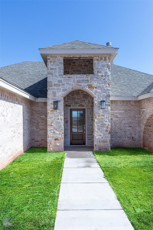 entrance to property featuring a shingled roof, stone siding, brick siding, and a lawn