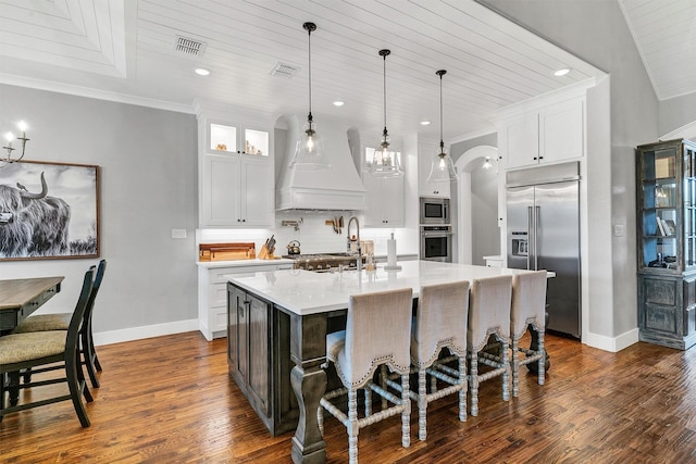 kitchen featuring white cabinets, custom exhaust hood, a kitchen bar, built in appliances, and an island with sink