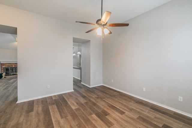 spare room featuring dark wood-style floors, a brick fireplace, a ceiling fan, and baseboards