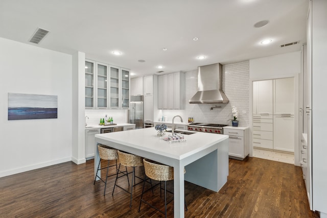 kitchen featuring white cabinets, wall chimney range hood, stainless steel appliances, a center island with sink, and dark hardwood / wood-style flooring