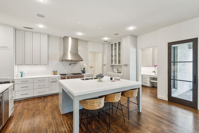 kitchen featuring a kitchen breakfast bar, dark wood-type flooring, white cabinets, a kitchen island with sink, and wall chimney range hood