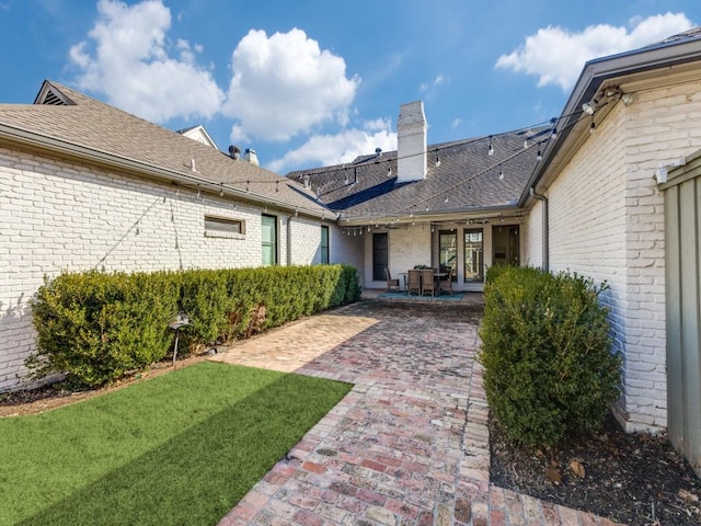 back of property with a shingled roof, a chimney, a yard, a patio area, and brick siding