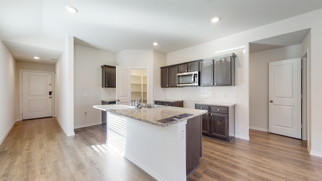 kitchen with sink, an island with sink, light hardwood / wood-style floors, dark brown cabinetry, and light stone counters