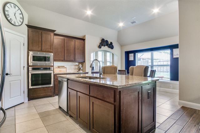 kitchen featuring appliances with stainless steel finishes, light stone countertops, dark brown cabinets, a center island with sink, and sink