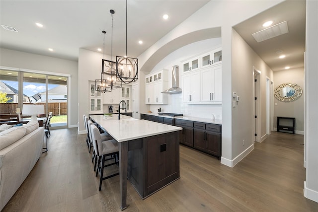 kitchen featuring white cabinetry, decorative light fixtures, stainless steel gas stovetop, a kitchen island with sink, and wall chimney range hood