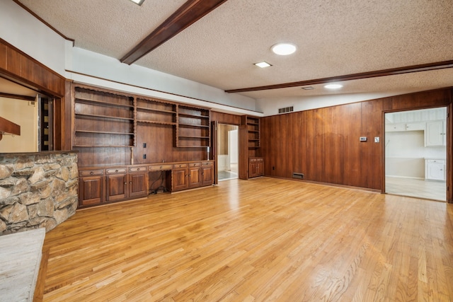 unfurnished living room with wood walls, a textured ceiling, built in desk, lofted ceiling with beams, and light wood-type flooring