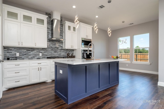 kitchen featuring hanging light fixtures, white cabinets, a kitchen island with sink, and wall chimney range hood