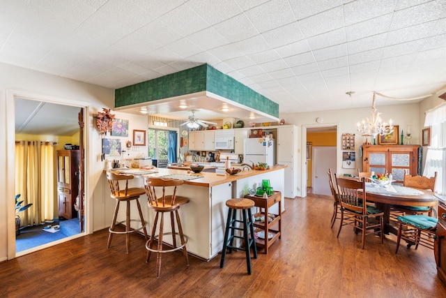 kitchen with kitchen peninsula, white appliances, a kitchen bar, ceiling fan with notable chandelier, and dark hardwood / wood-style flooring