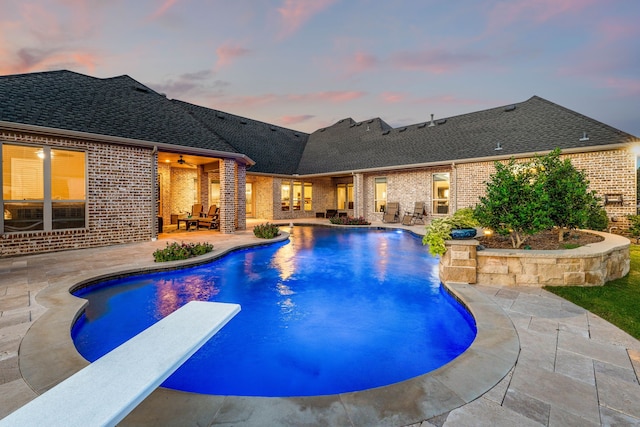 pool at dusk with a diving board, a patio area, and ceiling fan