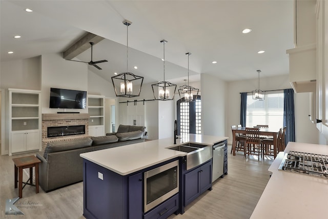 kitchen featuring a kitchen island with sink, stainless steel appliances, a fireplace, lofted ceiling with beams, and ceiling fan with notable chandelier