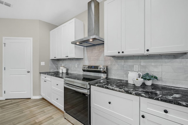 kitchen featuring dark stone counters, white cabinets, wall chimney exhaust hood, light wood-type flooring, and stainless steel electric range oven