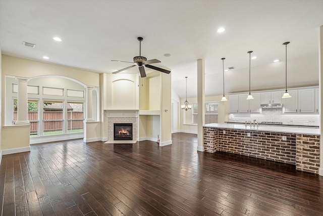 unfurnished living room featuring dark wood-type flooring, sink, ceiling fan with notable chandelier, decorative columns, and a brick fireplace