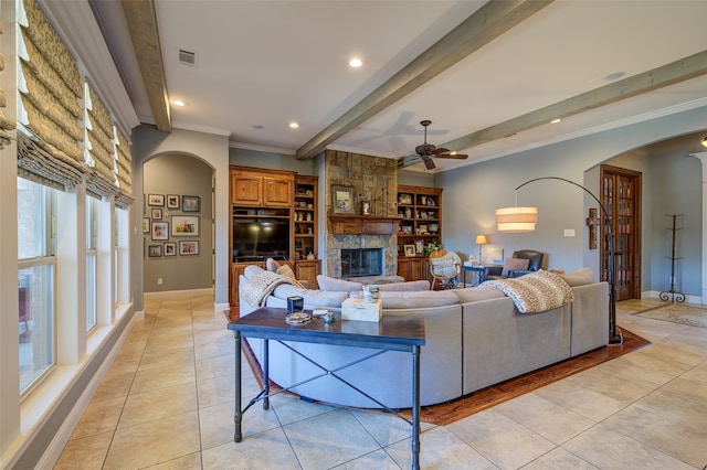 tiled living room featuring crown molding, a stone fireplace, beam ceiling, and ceiling fan