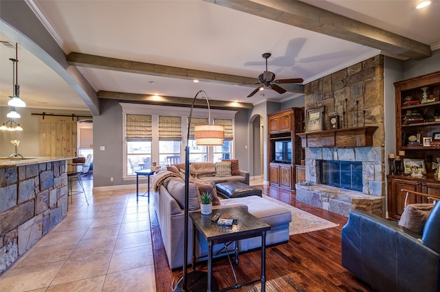 living room with ceiling fan, a stone fireplace, light wood-type flooring, crown molding, and a barn door