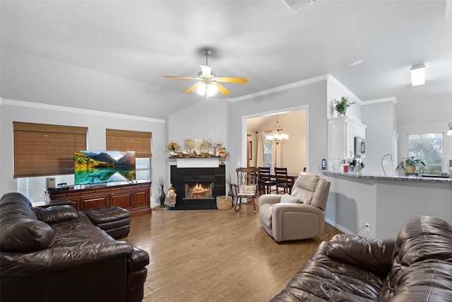 living room featuring ornamental molding, light hardwood / wood-style flooring, a tiled fireplace, ceiling fan with notable chandelier, and vaulted ceiling