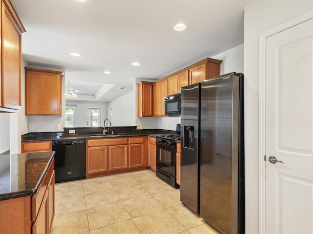 kitchen with dark stone counters, black appliances, sink, light tile patterned floors, and a tray ceiling