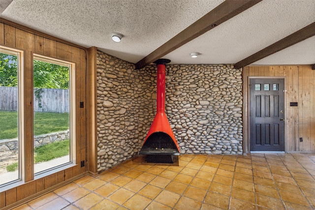 unfurnished living room with beamed ceiling, a textured ceiling, a wood stove, and wooden walls