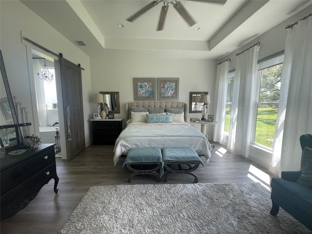 bedroom with ceiling fan, a barn door, dark wood-type flooring, and a tray ceiling