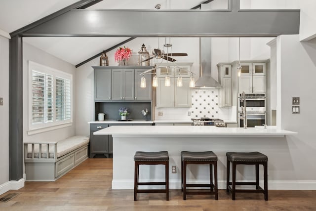 kitchen with wall chimney range hood, gray cabinets, light wood-type flooring, appliances with stainless steel finishes, and decorative light fixtures