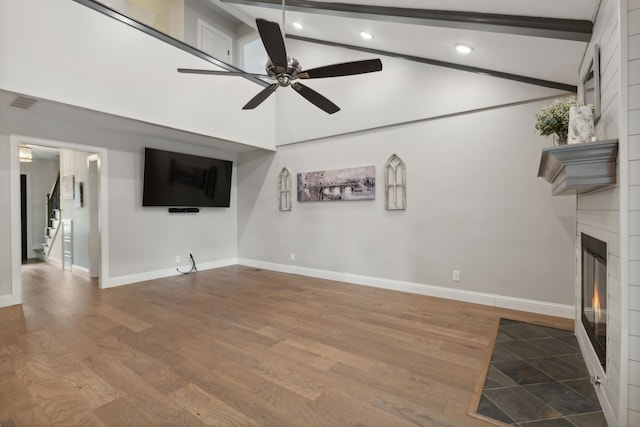 unfurnished living room with ceiling fan, beam ceiling, wood-type flooring, high vaulted ceiling, and a fireplace