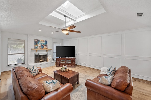 living room featuring light hardwood / wood-style floors, a textured ceiling, a brick fireplace, a skylight, and ceiling fan