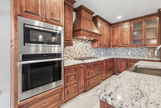 kitchen featuring custom range hood, backsplash, light stone countertops, light tile patterned floors, and stainless steel appliances