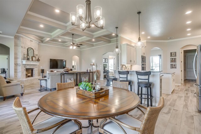 dining area with coffered ceiling, ceiling fan with notable chandelier, a stone fireplace, crown molding, and beam ceiling