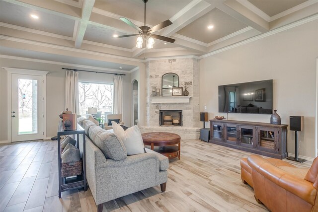 living room featuring ceiling fan, beamed ceiling, a stone fireplace, coffered ceiling, and crown molding