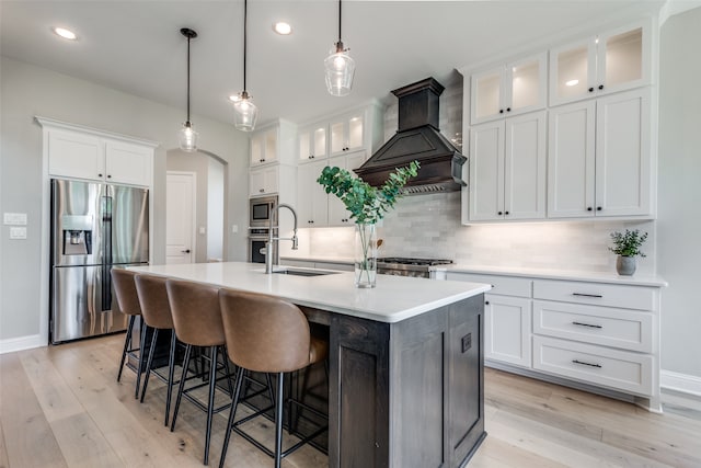 kitchen with premium range hood, white cabinets, a center island with sink, light hardwood / wood-style flooring, and appliances with stainless steel finishes