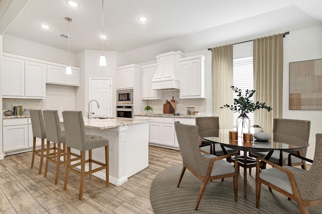 kitchen with premium range hood, stainless steel appliances, decorative light fixtures, a center island with sink, and white cabinetry