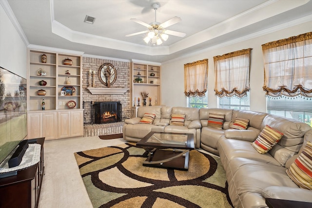 carpeted living room featuring a brick fireplace, built in features, ceiling fan, crown molding, and a raised ceiling