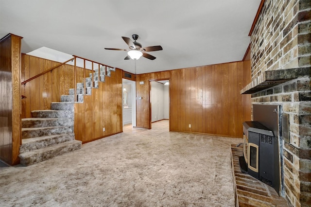 unfurnished living room featuring ceiling fan, light carpet, wooden walls, and a wood stove