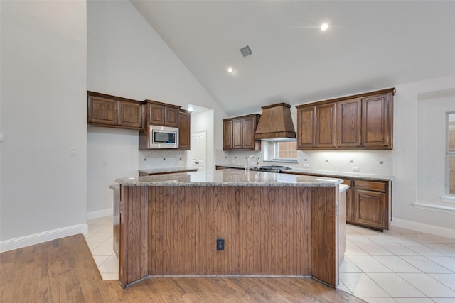 kitchen with stainless steel microwave, premium range hood, a kitchen island with sink, high vaulted ceiling, and light stone countertops