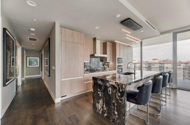 interior space with dark wood-type flooring, backsplash, wall chimney exhaust hood, dark stone counters, and a kitchen island with sink