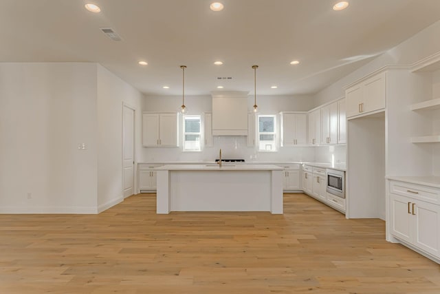 kitchen with hanging light fixtures, stainless steel microwave, an island with sink, custom range hood, and white cabinets