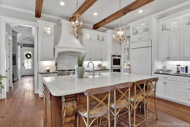 kitchen with beam ceiling, decorative light fixtures, a kitchen island with sink, a chandelier, and white cabinetry