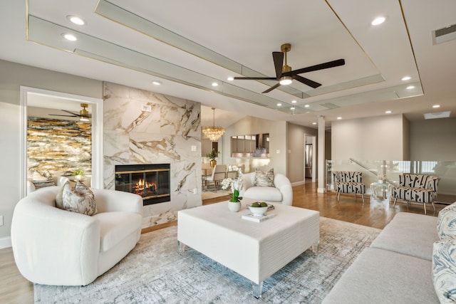 living room with ceiling fan with notable chandelier, light wood-type flooring, a tray ceiling, and a fireplace