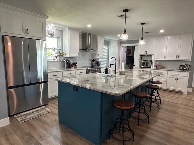 kitchen featuring stainless steel appliances, a center island with sink, wall chimney exhaust hood, and white cabinetry