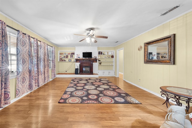 living room featuring ceiling fan, built in shelves, a brick fireplace, crown molding, and light hardwood / wood-style floors