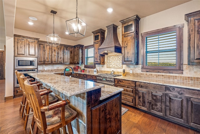 kitchen with premium range hood, a center island with sink, appliances with stainless steel finishes, an inviting chandelier, and light stone countertops