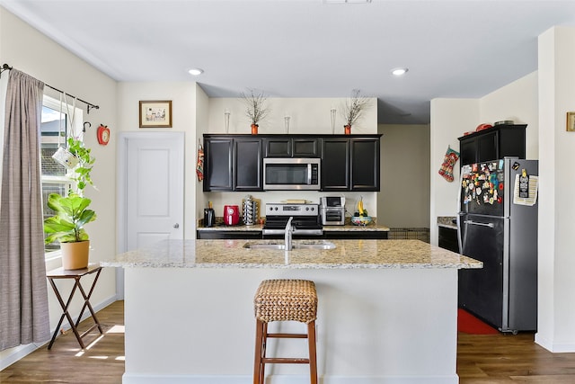 kitchen featuring appliances with stainless steel finishes, an island with sink, light stone countertops, dark hardwood / wood-style flooring, and sink