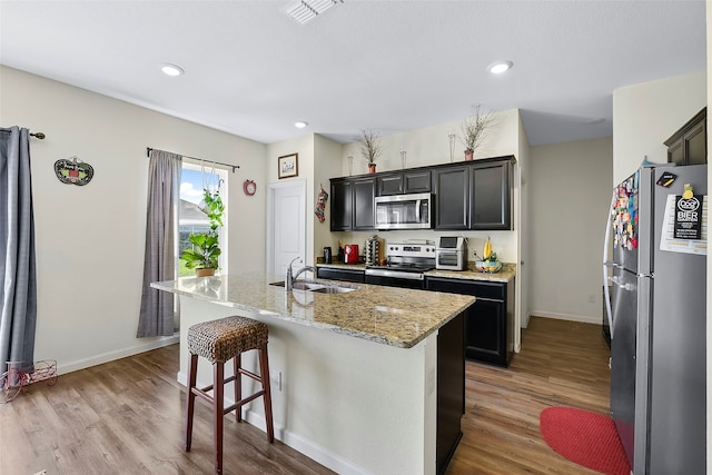 kitchen with light stone countertops, light wood-type flooring, appliances with stainless steel finishes, and a kitchen island with sink