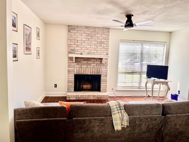 living room featuring baseboards, ceiling fan, wood finished floors, a textured ceiling, and a brick fireplace