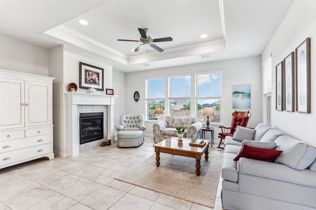 living room featuring a tray ceiling, a tiled fireplace, light tile patterned floors, crown molding, and ceiling fan