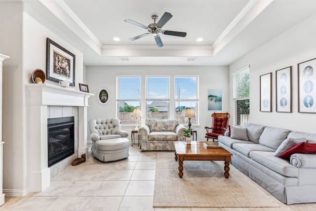 living room with ceiling fan, a tray ceiling, a tiled fireplace, and ornamental molding