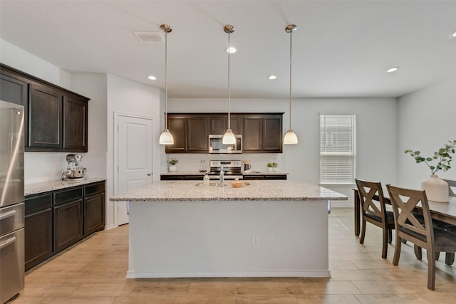 kitchen with hanging light fixtures, stainless steel appliances, light stone counters, an island with sink, and dark brown cabinets